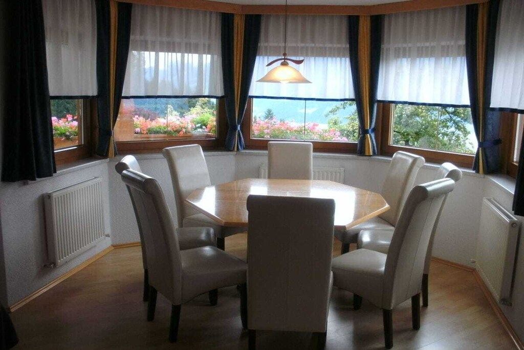 Ein Esszimmer mit einem runden Tisch und sechs Stühlen, großen Fenstern mit Vorhängen und Blick ins Grüne.