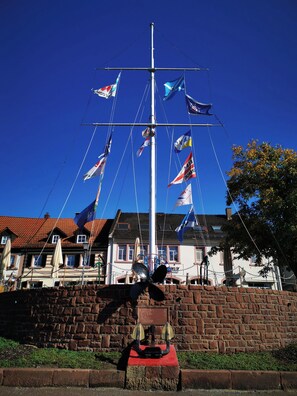 A tall flagpole with multiple flags, a brick wall, and a building with a red-tiled roof in the background. - Charming 1-Bedroom Apartment in Historic Marktheidenfeld (Marktheidenfeld)