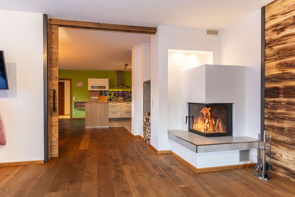 A modern living room with a fireplace, wooden flooring, and a kitchen in the background.
