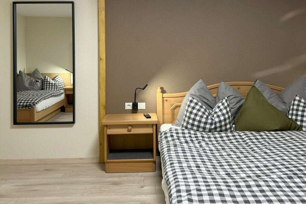 A bedroom with a checkered bedspread, a nightstand with a lamp, and a mirror on the wall.