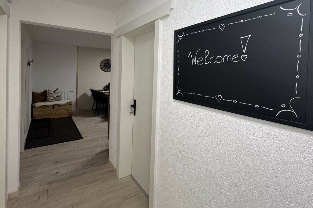 A hallway with a blackboard displaying 'Welcome' and a door leading to a room with a bed and a chair.