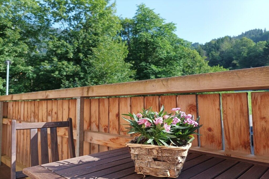 A wooden deck with a table and a chair, a woven basket with pink flowers, and a wooden fence with greenery in the background.