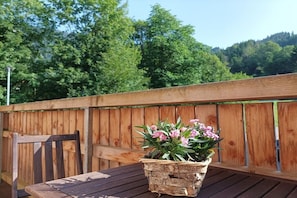 Een houten terras met een tafel en stoel, een gevlochten mand met roze bloemen, en een houten omheining met beplanting op de achtergrond.