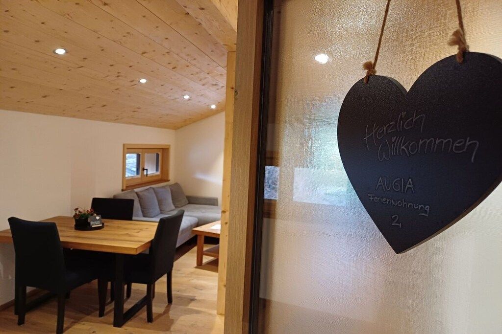 Quarto com teto de madeira, mesa de jantar com cadeiras, um sofá e um letreiro em formato de coração com a mensagem 'Herzlich Willkommen' e 'AUGIA Ferienwohnung 2'.