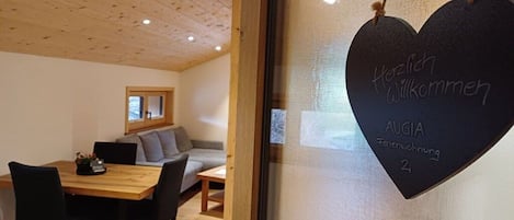 A room with a wooden ceiling, a dining table with chairs, a sofa, and a heart-shaped sign with 'Herzlich Willkommen' and 'AUGIA Ferienwohnung 2' written on it.