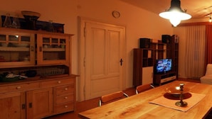 Zona pranzo con tavolo in legno, sedie, un mobile con ante in vetro, una televisione e un orologio appeso al muro.