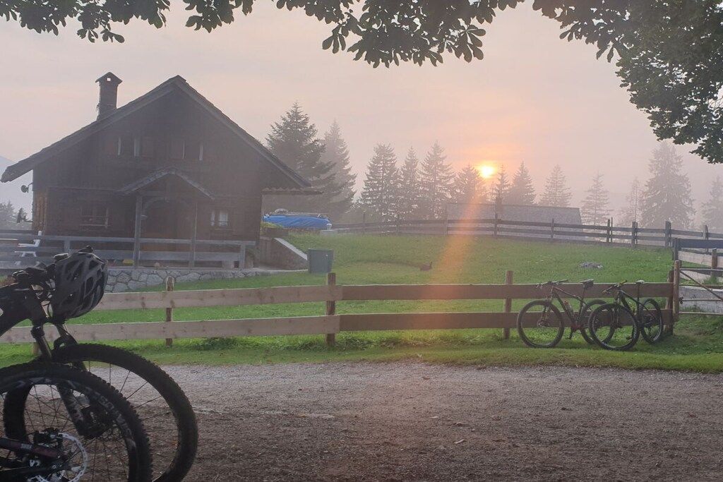 Une cabane en bois rustique, accessible par un chemin de terre, entourée d’une clôture en bois et de vélos.