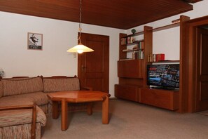 Ein Wohnzimmer mit einem Holzsofa, einem Holztisch, einem Fernseher auf einem Holzständer und einer Hängeleuchte.