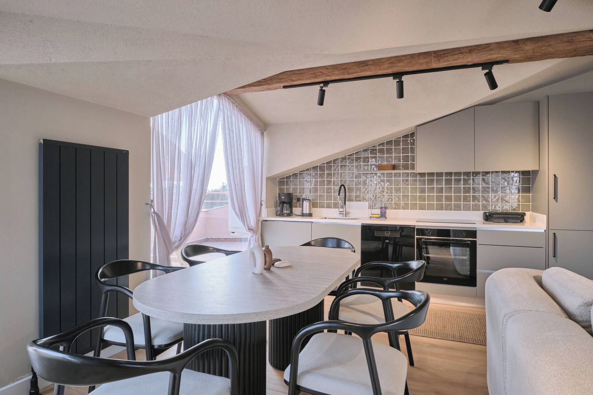 Una cocina moderna con horno empotrado, una zona de comedor con una mesa redonda y sillas, y vistas al exterior a través de una ventana con cortinas translúcidas.