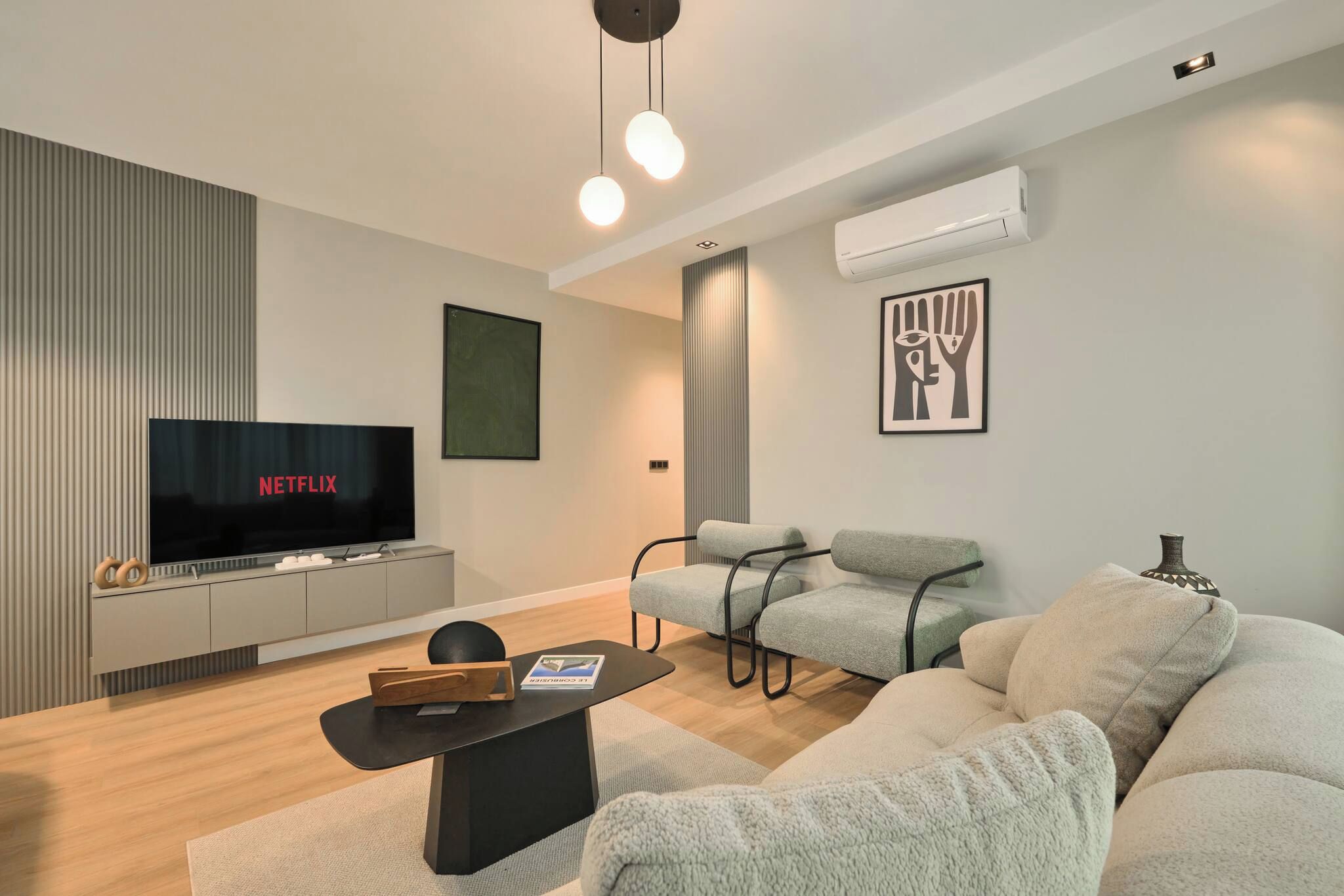 A modern living room with a flat-screen TV, grey sofas, a coffee table, and a wall-mounted air conditioner.