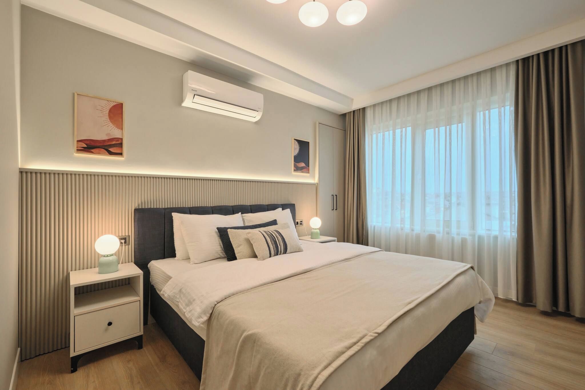 A modern hotel room with a large bed, bedside lamps, an air conditioning unit, and a window with curtains.
