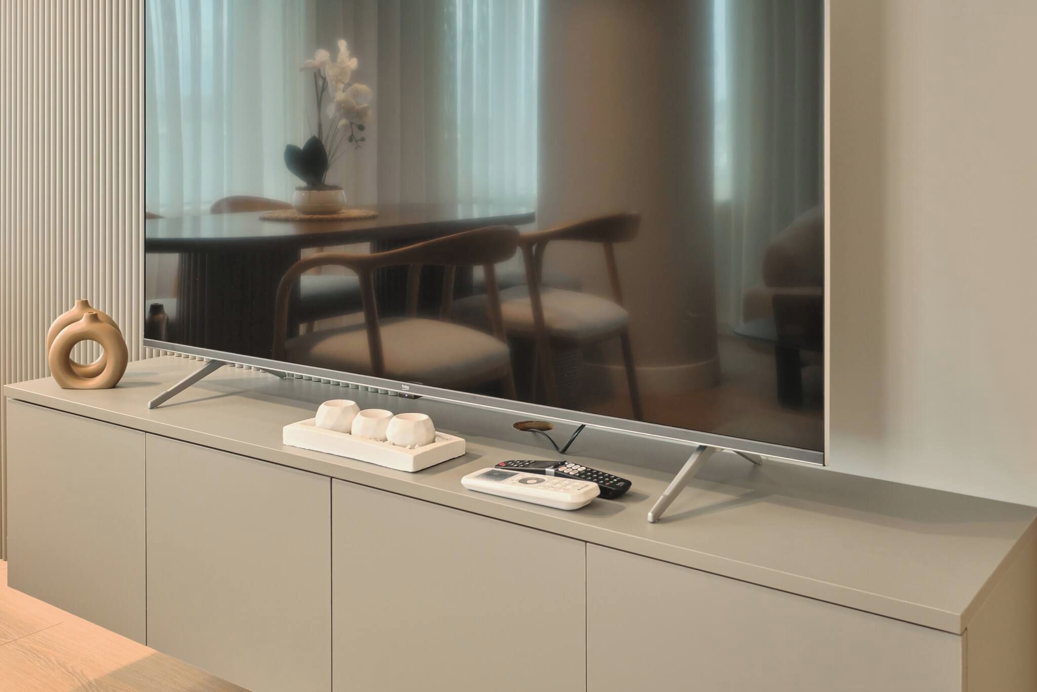 Uma sala de estar moderna com uma televisão de tela plana grande fixada na parede, um vaso de madeira, um tabuleiro com velas e um controle remoto sobre um móvel.