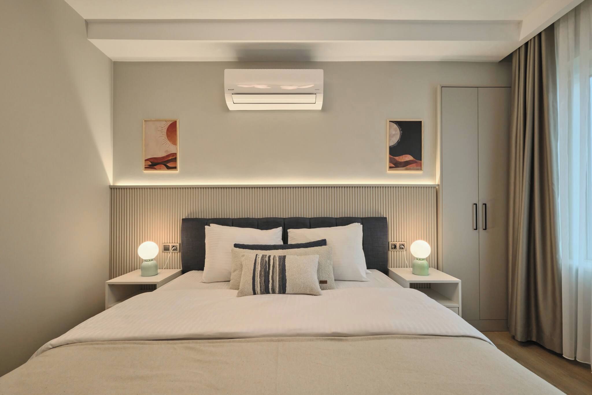 A modern hotel room with a large bed, two bedside tables with lamps, a wall-mounted air conditioner, and artwork above the bed.
