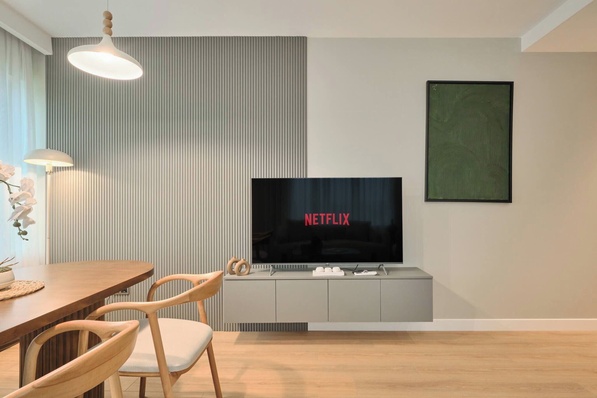 A modern living room with a flat-screen TV displaying 'NETFLIX,' a dining area with a wooden table and chairs, and a large abstract painting on the wall.