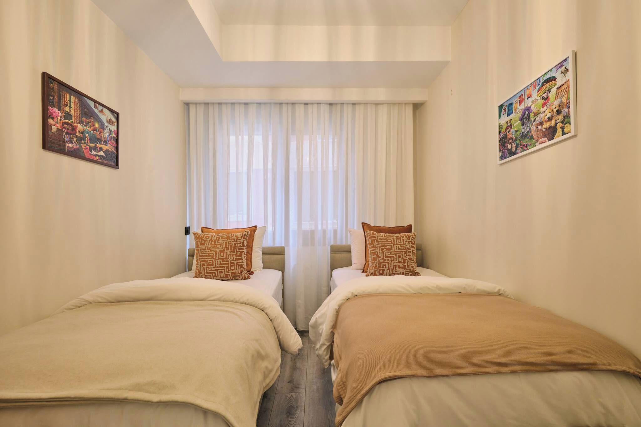 Habitación de hotel con dos camas individuales, un ventanal con cortinas translúcidas y dos cuadros enmarcados en las paredes.