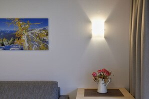 Une pièce avec un tableau représentant un paysage de montagne, une table de chevet ornée d’un vase de fleurs et un luminaire fixé au mur.