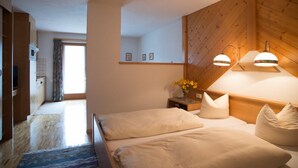 Een slaapkamer met een houten hoofdeinde, twee bedden, een nachtkastje met een bloemenvaas, en uitzicht op de keuken.