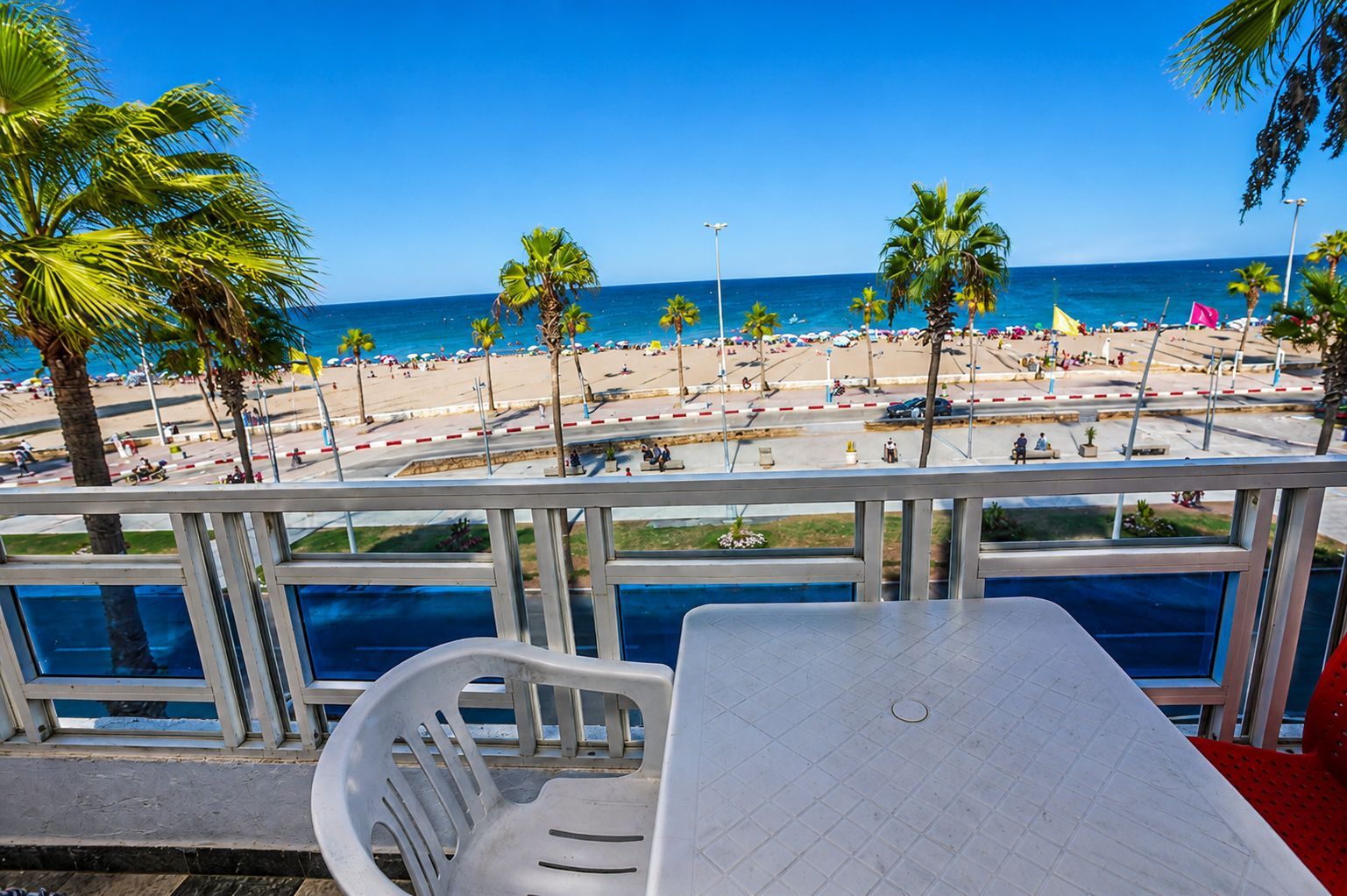 A beach view from a balcony with a table and chairs, palm trees, and a clear blue sky.