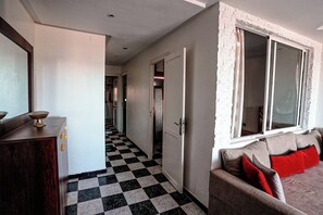 Ein Korridor mit Schachbrettmuster führt zu einem Zimmer mit großem Fenster und einer Couch mit roten Kissen.