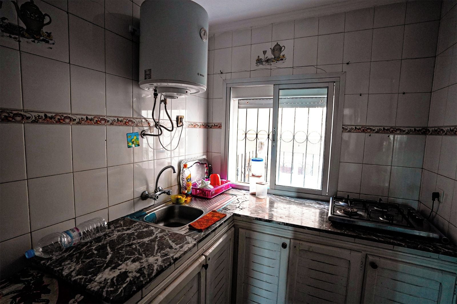 A kitchen with white tiled walls, a sink, a stove, and a window with a view.