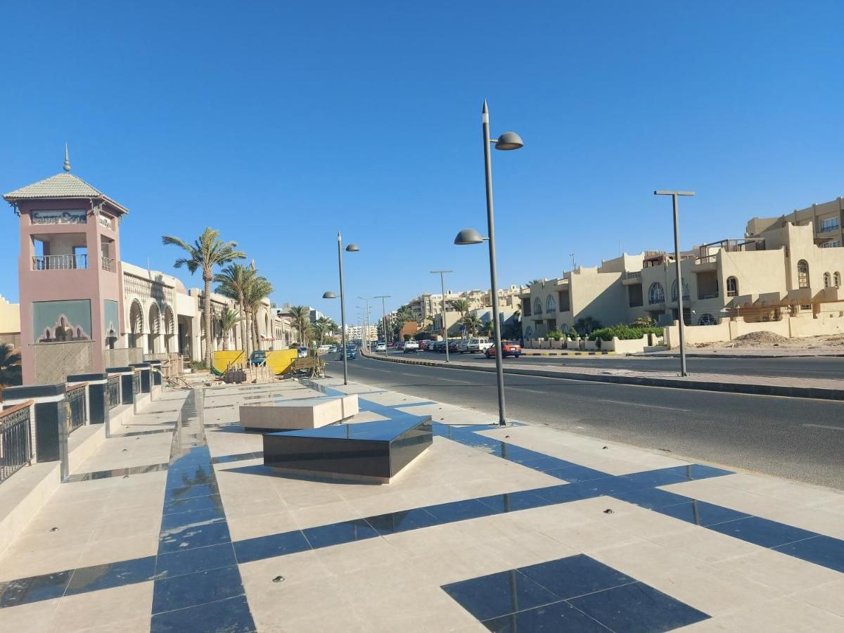 Une large avenue urbaine bordée d’une place moderne, de palmiers et d’immeubles à l’architecture traditionnelle et contemporaine.