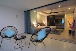 Een moderne woonruimte met een glazen scheidingswand, een bank, een bed en twee rieten fauteuils met kussens.