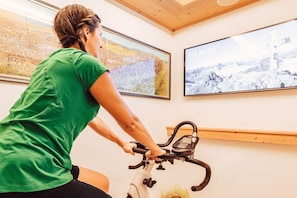 Une personne fait de l’exercice sur un vélo d’appartement dans une pièce dotée d’une grande fenêtre et d’un tableau encadré accroché au mur.