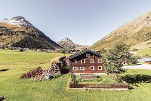 A two-story wooden chalet with a sloped roof, surrounded by a grassy field and mountains in the background. - Apart Zimalis Galtür Ischgl 3-5 Per. (Galtür)