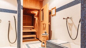 Een houten sauna met een bankje en een klein krukje, een witte badkuip met een zwart mozaïekrand, en een douchegedeelte met een douchekop.