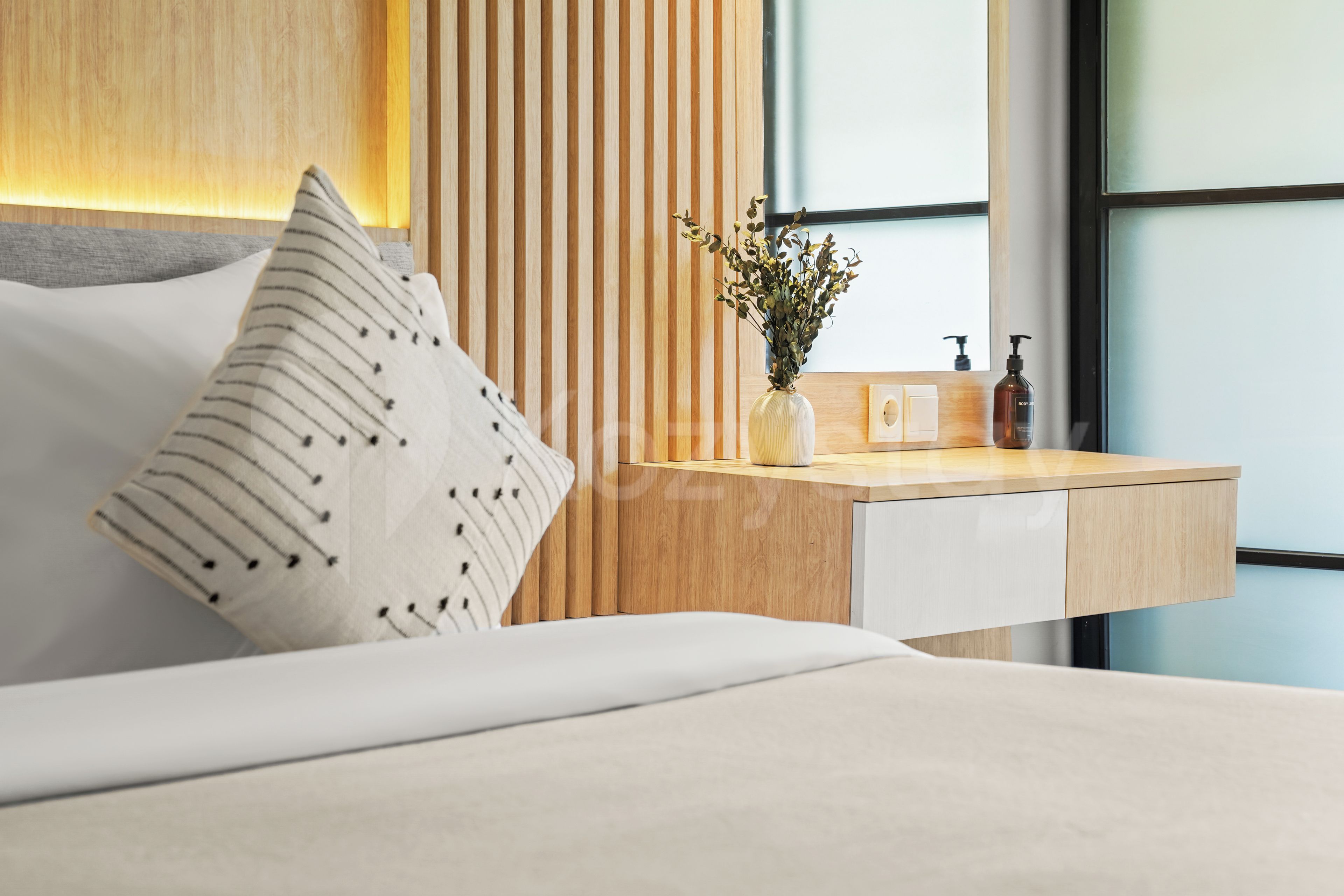 A modern hotel room with a bed, a wooden bedside table, a vase with dried flowers, a bottle of body lotion, and a window with a view.