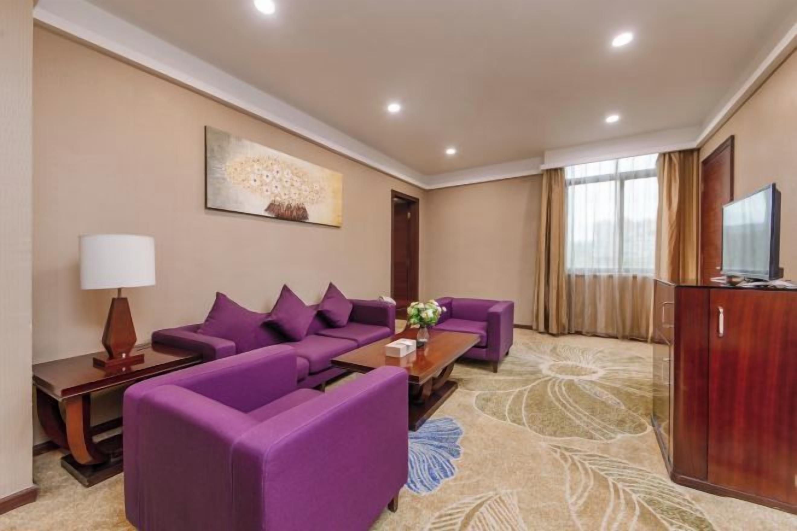 A hotel room with a purple sofa set, a flat-screen TV, and a large window with curtains.