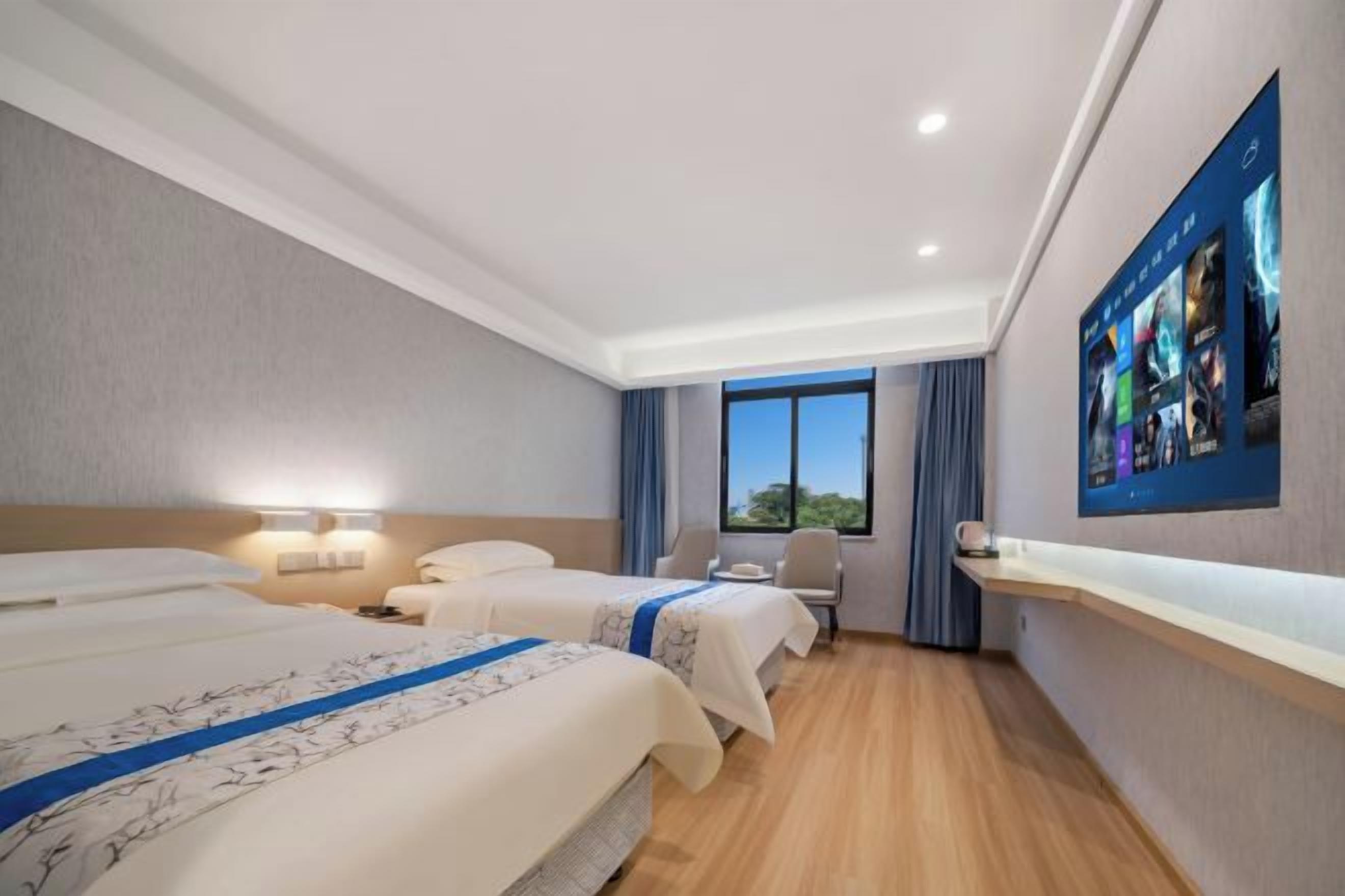 Une chambre d’hôtel avec deux lits, un bureau, une chaise, une fenêtre avec des rideaux et un écran fixé au mur qui affiche des images.