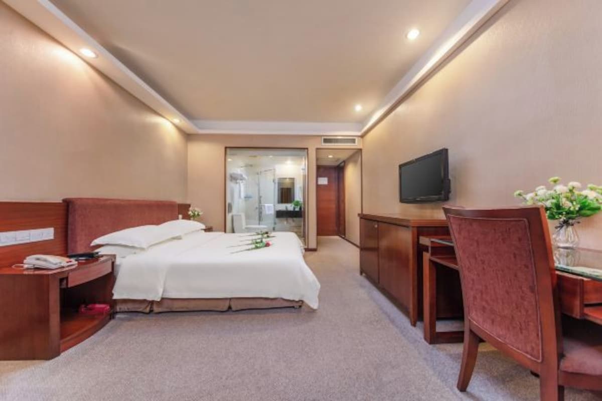 Habitación de hotel con cama, mesita de noche, silla, escritorio, televisor y una pequeña mesa con un jarrón de flores.
