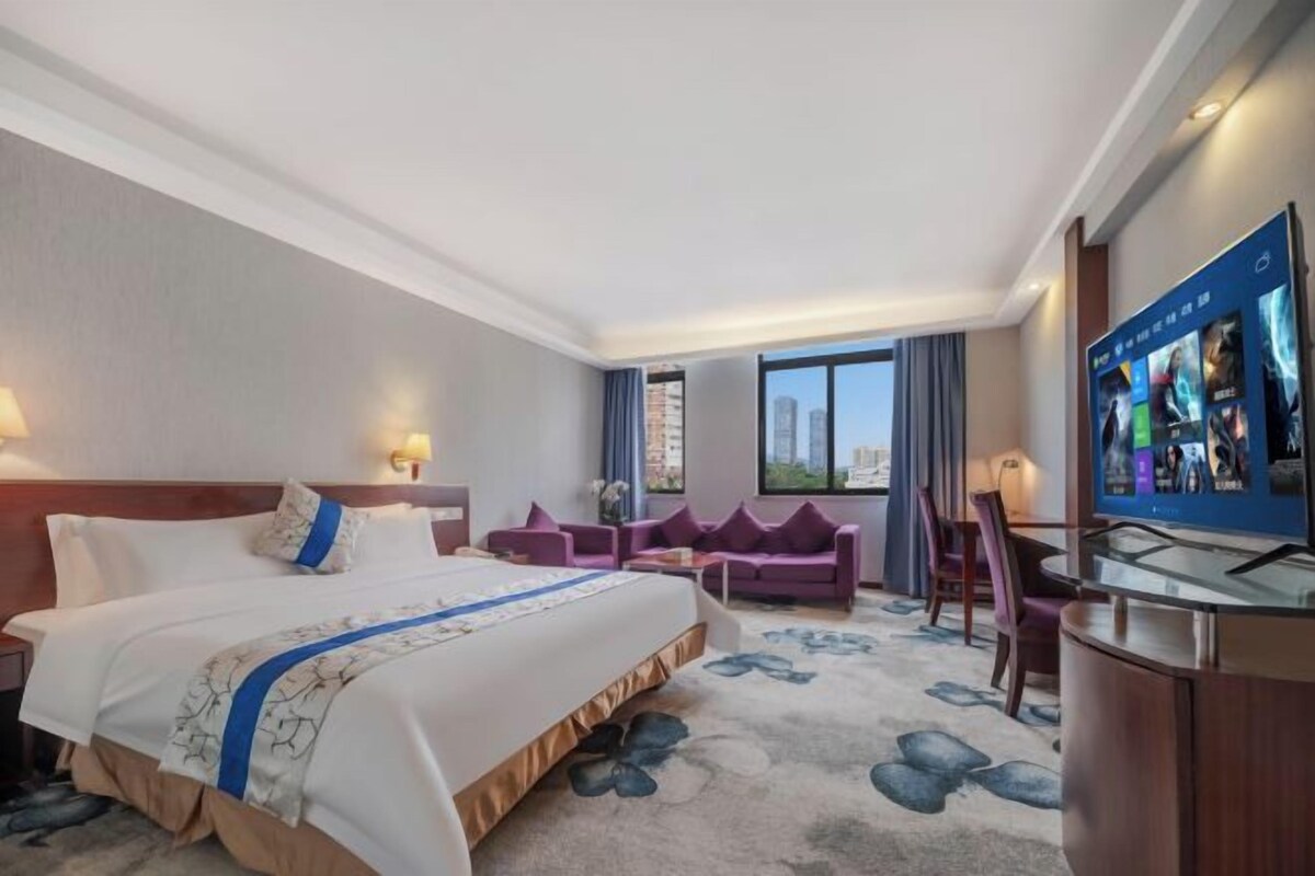 Habitación de hotel con una cama grande, un escritorio con televisor de pantalla plana, una zona de estar con sillas y vistas a la ciudad.