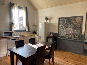 Kompakti keittiö, jossa on mikroaaltouuni, jääkaappi ja pesuallas. Keittiössä on ruokapöytä tuoleineen sekä suuri ikkuna, jossa on verhot.