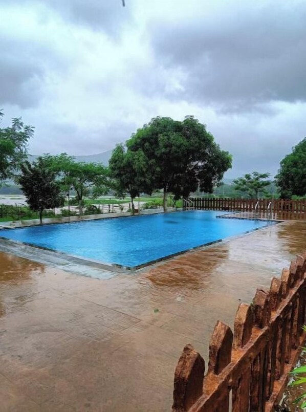 A rectangular swimming pool with a wooden fence, surrounded by trees and a cloudy sky.