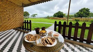 A covered outdoor seating area with a table set for a meal, surrounded by a wooden fence and a view of a grassy field.