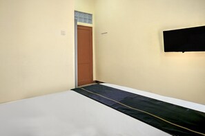 Ein schmaler Raum mit einem weißen Bett, einer braunen Tür und einem an der Wand befestigten Fernseher.