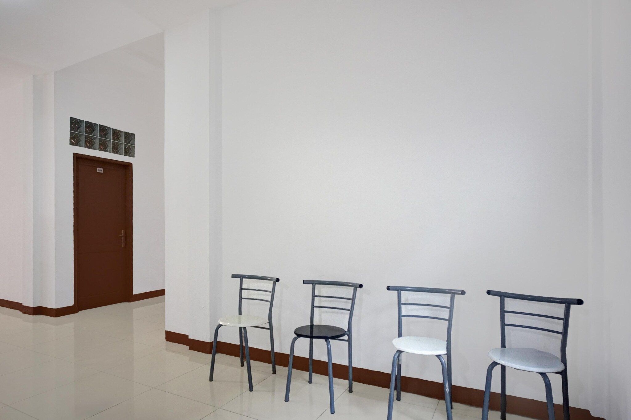 Un pasillo con tres sillas alineadas contra una pared, una puerta con un número y un ventanal con diseño.