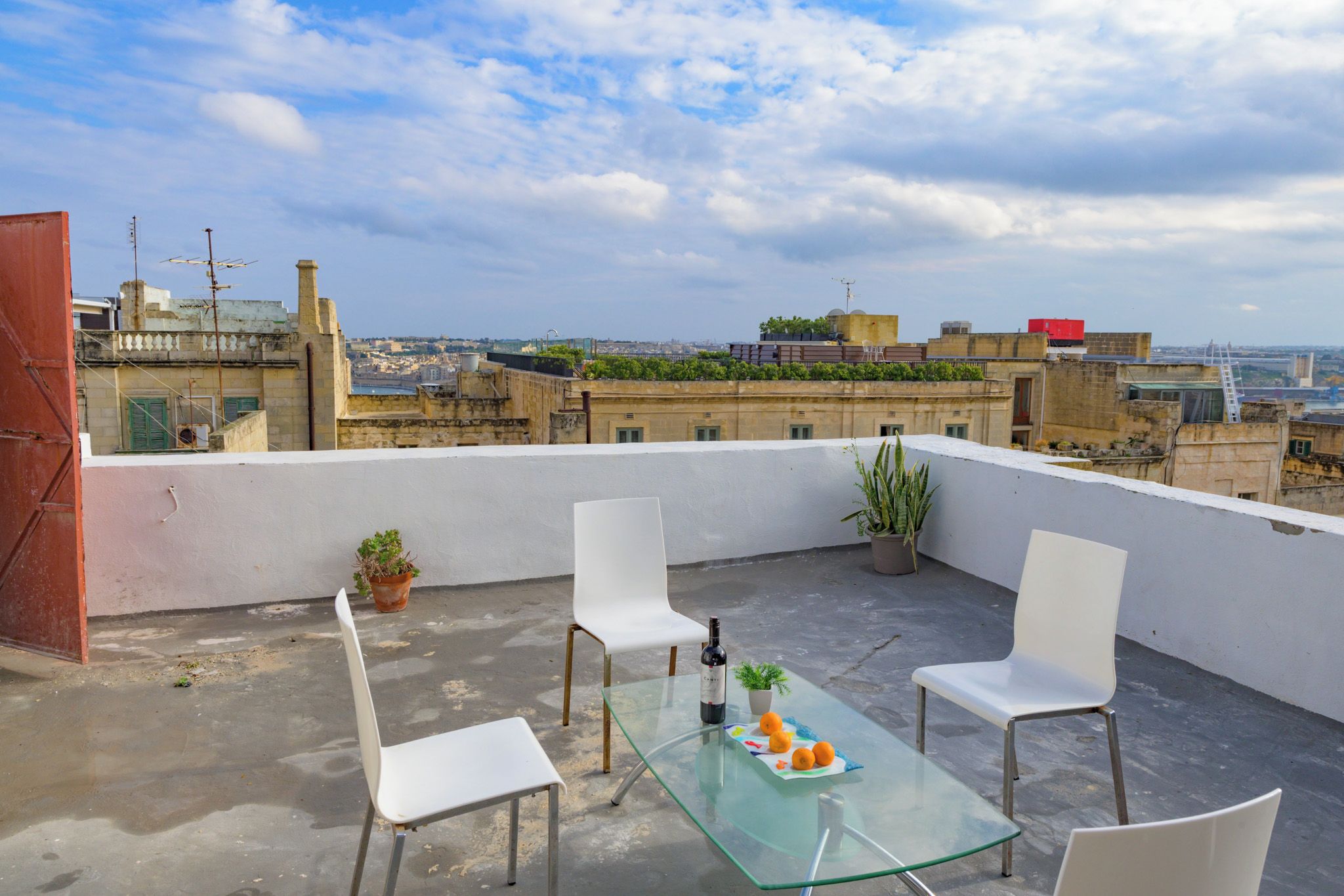 Una terraza en la azotea con una mesa de vidrio, sillas blancas, una botella de vino y una planta en maceta.