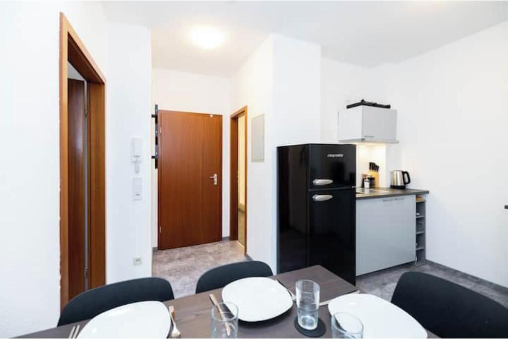 A compact kitchen with a refrigerator, stove, and coffee maker. There is a dining table with chairs and a door leading to another room.