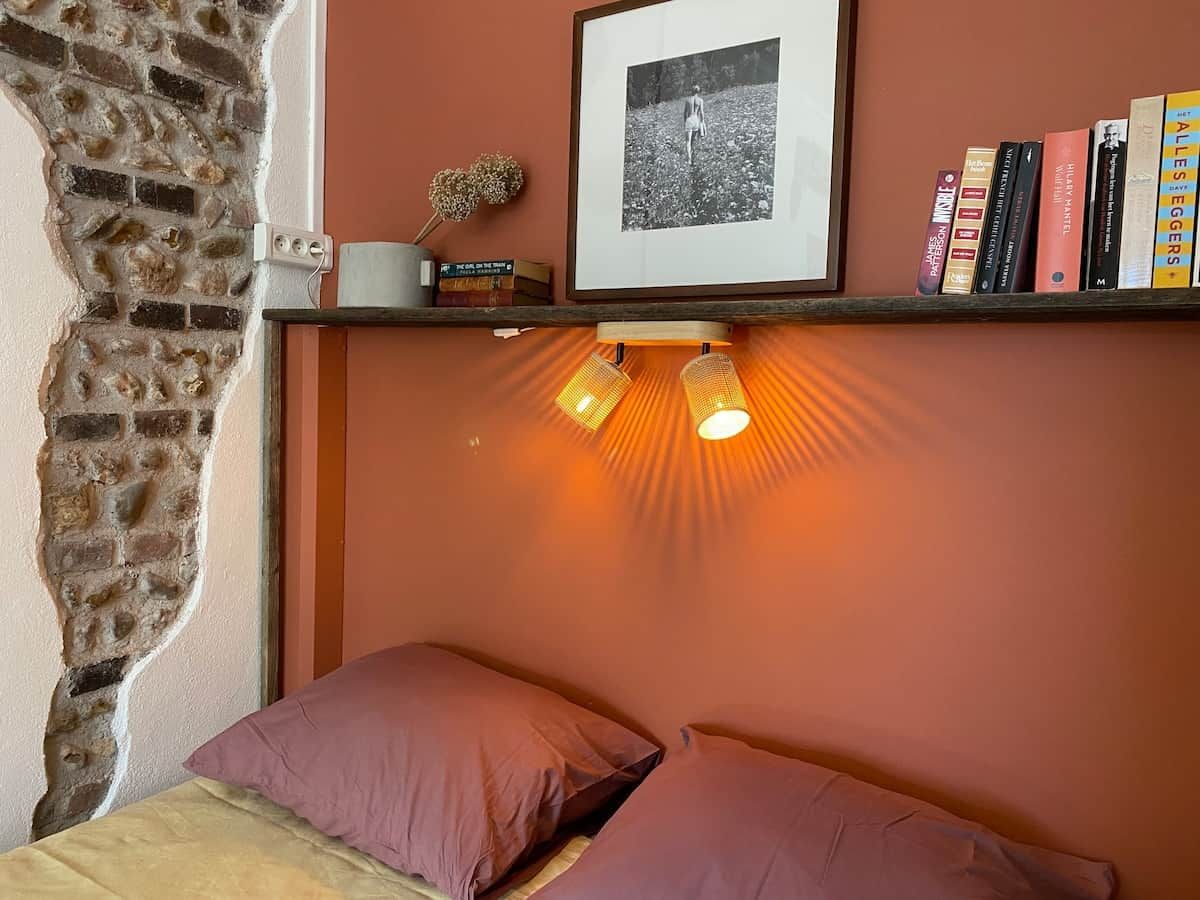Un dormitorio con pared de ladrillo, una cama con dos almohadas, una estantería con libros y una foto enmarco, y un aplique de pared.