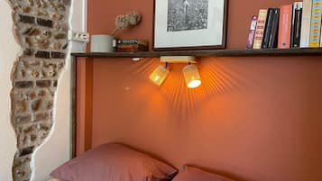 Un dormitorio con pared de ladrillo, una cama con dos almohadas, una estantería con libros y una foto enmarco, y un aplique de pared.