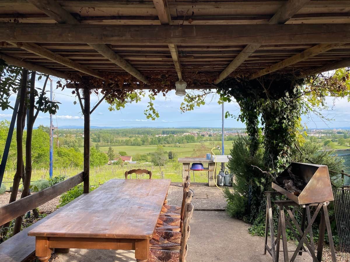 Eine Terrasse im Freien mit Holztisch und -stühlen, einem Grill und Blick auf eine grüne Landschaft.