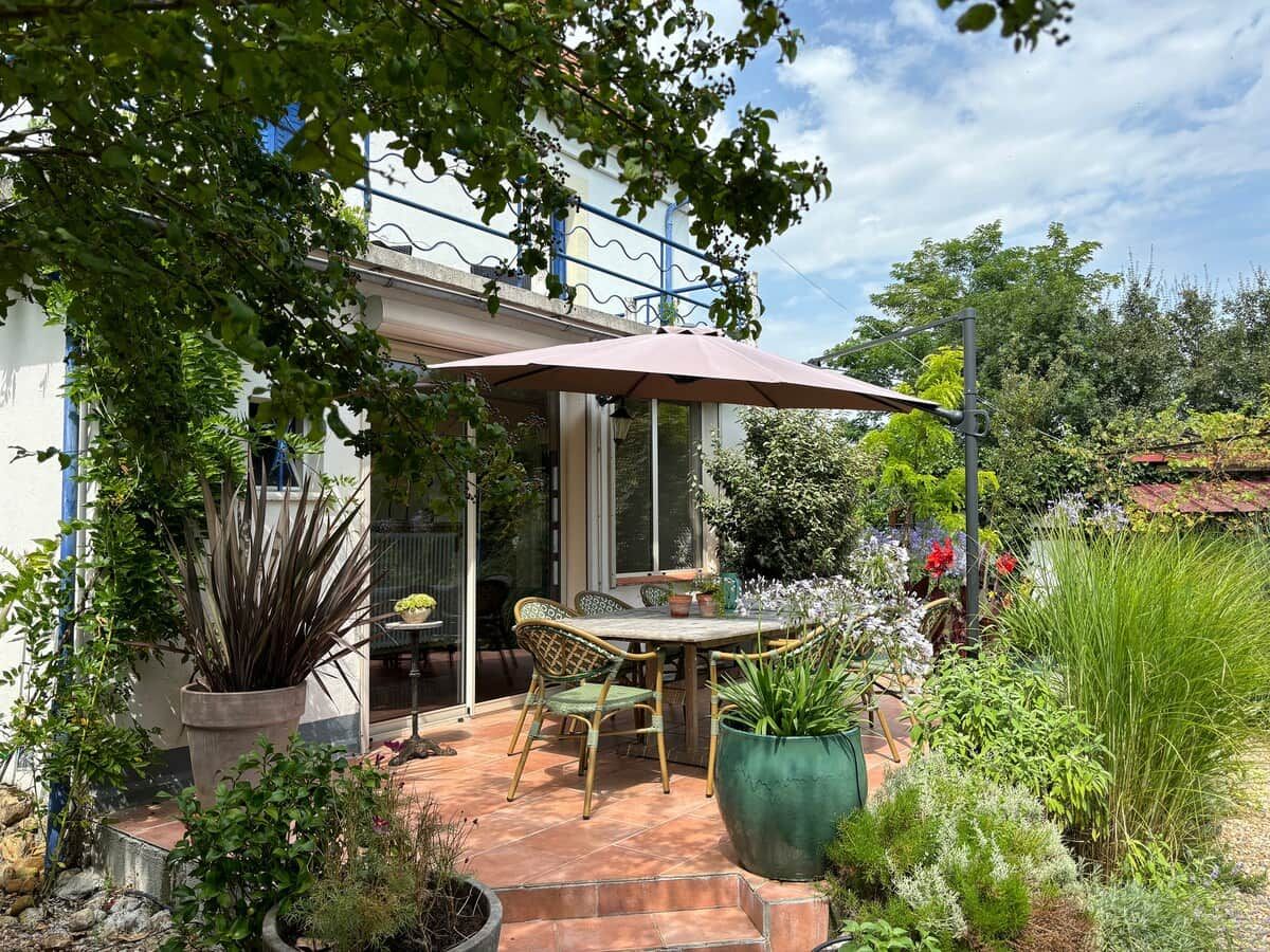 Une terrasse avec une table et des chaises, entourée de plantes en pot et d’un grand parasol.
