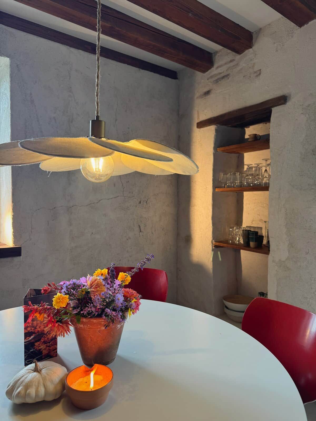 Een eetgedeelte met een ronde tafel, een hanglamp, een bloevas, een brandende kaars en een rode stoel.