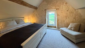 Ein Schlafzimmer mit schrägem Dach, einem Bett, einer Couch und einem Fenster mit Blick auf Grünflächen.
