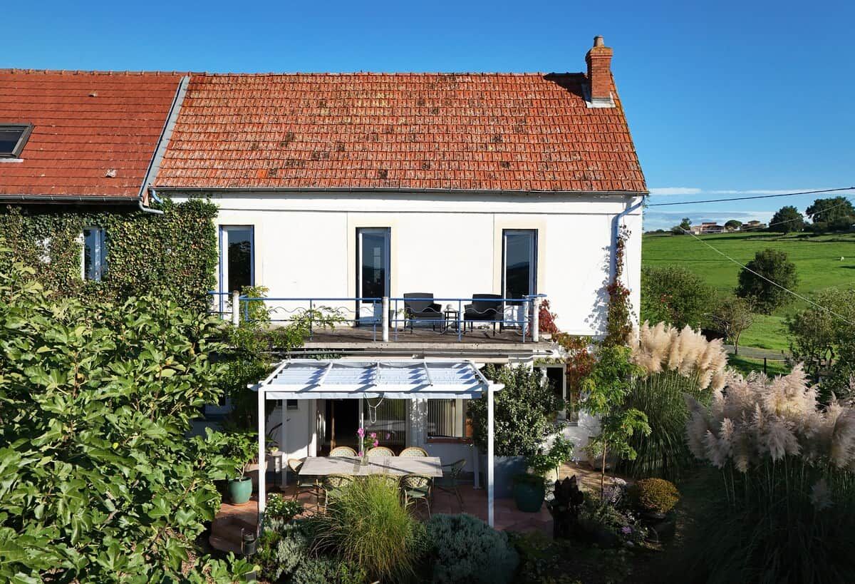 Ein zweistöckiges Haus mit rotem Ziegeldach, einer überdachten Terrasse mit Gartenmöbeln und einem Garten mit verschiedenen Pflanzen und Bäumen.