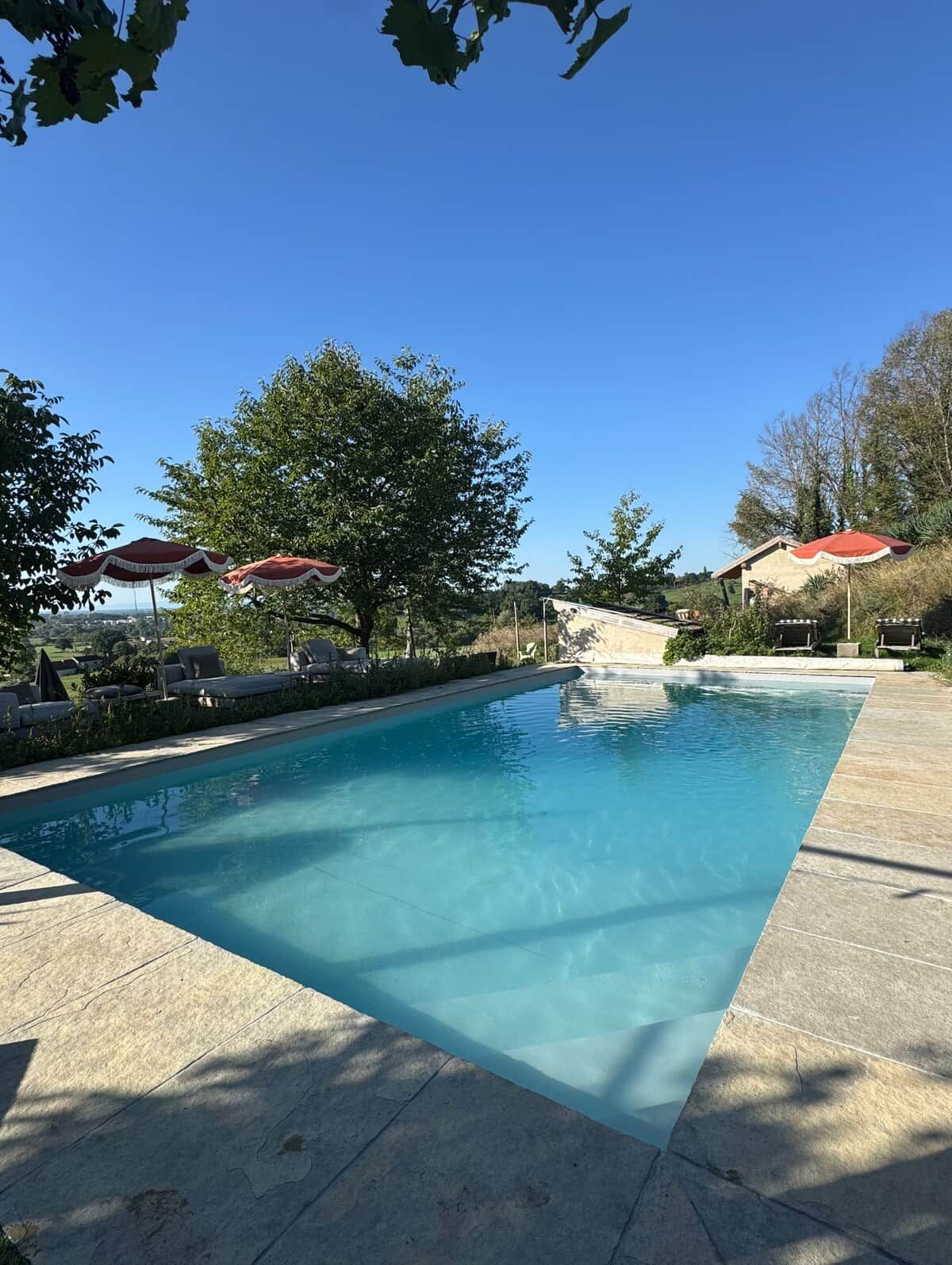 Una piscina con sillas de descanso y sombrillas, rodeada de árboles y un cielo despejado.