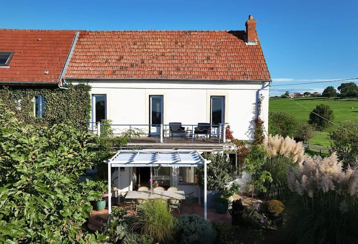 Une maison à deux étages avec un toit recouvert de tuiles rouges, une terrasse couverte avec des meubles d’extérieur et un jardin où poussent différentes espèces de plantes.
